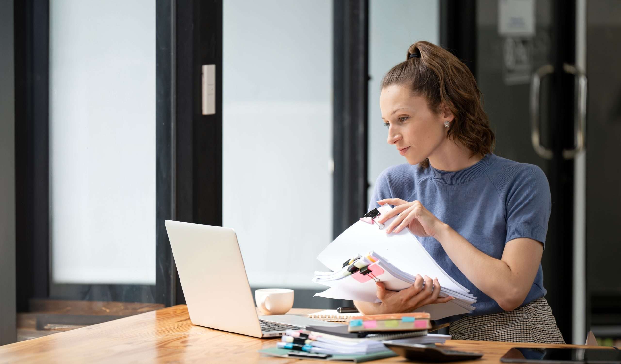 Young busy business woman or company employee holding accounting bookkeeping documents checking financial data or marketing report working in office with laptop. Paperwork management